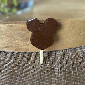 May include: A chocolate-covered ice cream bar shaped like a mouse's head, with a bite taken out of the top. The ice cream is on a wooden stick. The background is a wooden tray and a woven mat.
