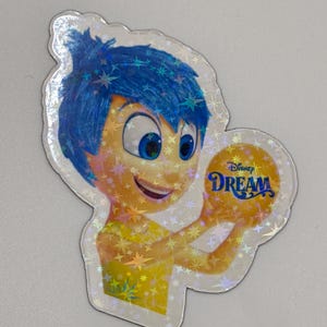 May include: A holographic sticker featuring the character Joy from the Disney movie Inside Out. Joy has blue hair and a yellow dress, holding a golden ball with the word "Dream" on it. The sticker has a star pattern.