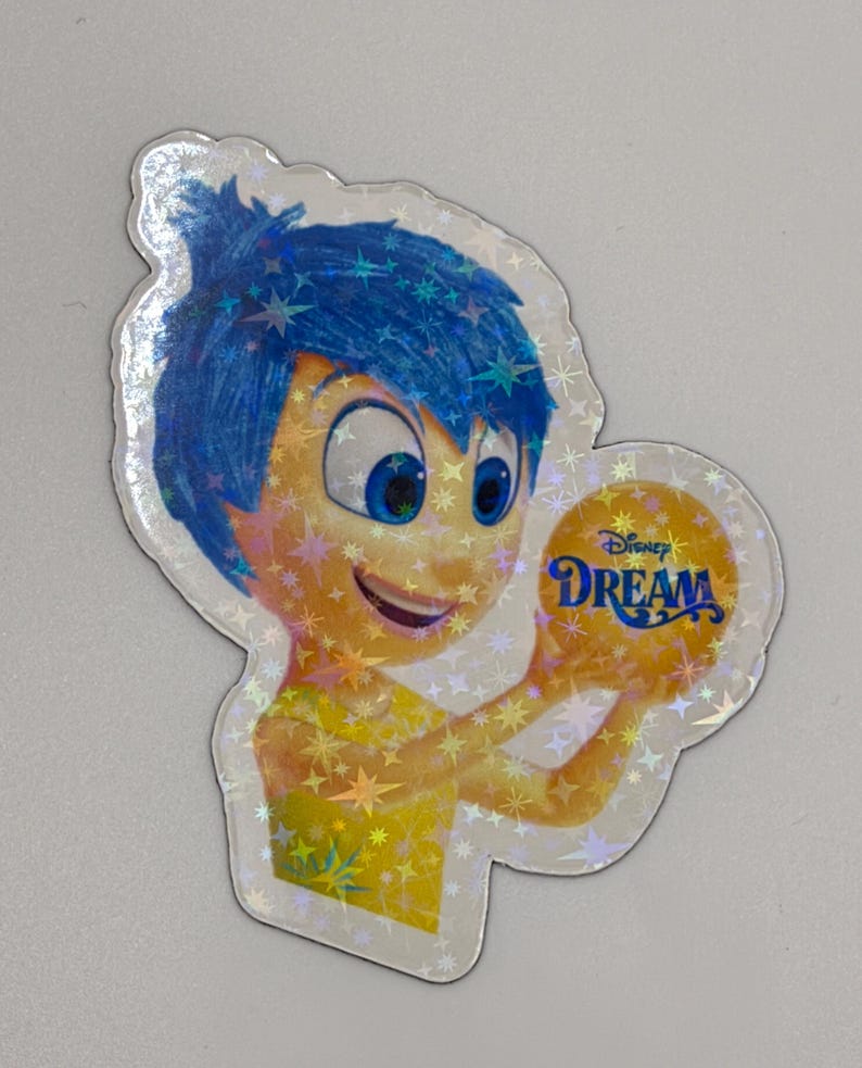 May include: A holographic sticker featuring the character Joy from the Disney movie Inside Out. Joy has blue hair and a yellow dress, holding a golden ball with the word "Dream" on it. The sticker has a star pattern.