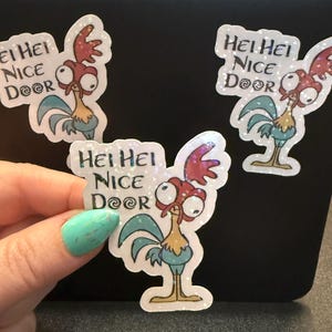 May include: Three glittery stickers featuring a cartoon rooster with the text "Hei Hei Nice Door". The rooster has red, blue, and yellow coloring. The stickers are on a black surface.