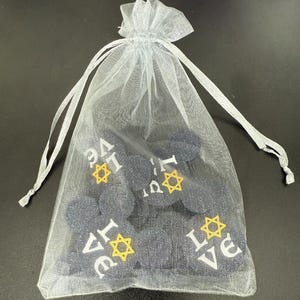 May include: A sheer white drawstring bag filled with dark blue, circular objects. The objects have the word "LOVE" and a Star of David symbol printed on them in white and yellow. The bag is tied with a white ribbon.