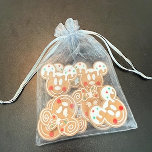 May include: A light blue organza bag filled with gingerbread-style Mickey Mouse and gingerbread man-shaped ornaments. The ornaments are brown with white icing and red, green, and white accents. The bag is tied with a matching ribbon.