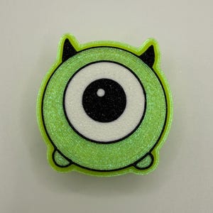 May include: A lime green, circular character with a large white eye, black pupil, and small black horns. The character has a textured, glittery appearance and a black outline. The design is reminiscent of a cartoon monster.