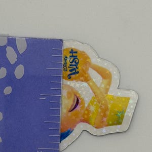 May include: A Disney Wish sticker featuring a character in a yellow dress holding a glowing orb. The sticker has a holographic finish and the Disney Wish logo is visible. The sticker is approximately 2 inches in height.