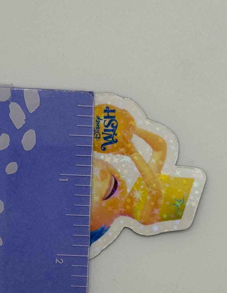 May include: A Disney Wish sticker featuring a character in a yellow dress holding a glowing orb. The sticker has a holographic finish and the Disney Wish logo is visible. The sticker is approximately 2 inches in height.