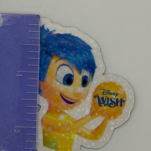 May include: A holographic sticker featuring the character Joy from the Disney movie "Inside Out." Joy has blue hair and is holding a yellow orb with the words "Disney Wish." The sticker is approximately 2 inches in height.