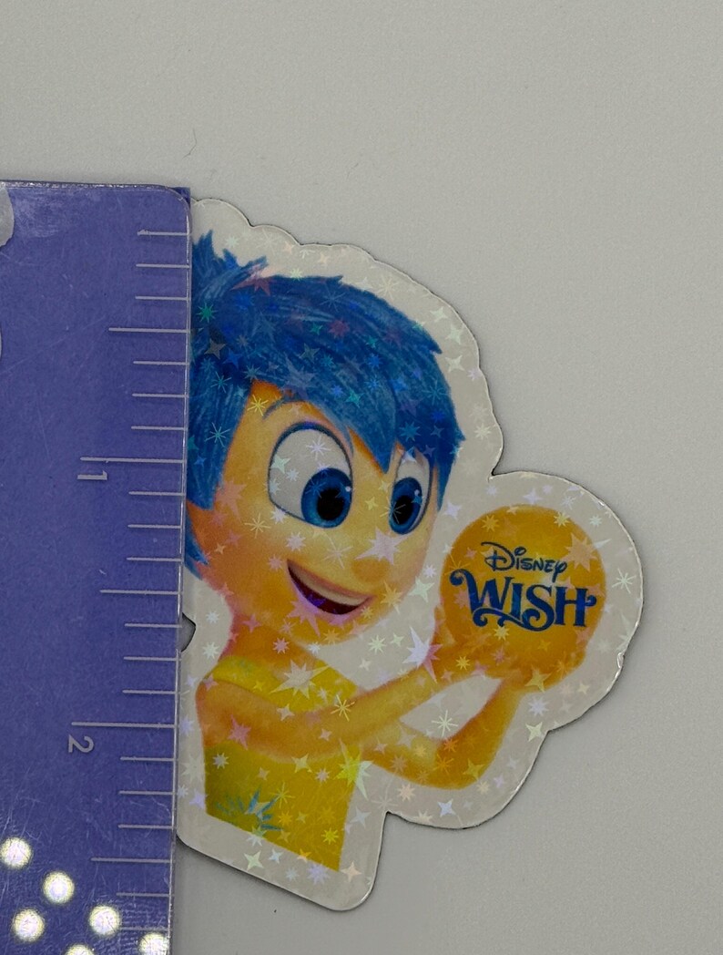 May include: A holographic sticker featuring the character Joy from the Disney movie "Inside Out." Joy has blue hair and is holding a yellow orb with the words "Disney Wish." The sticker is approximately 2 inches in height.