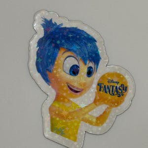 May include: A holographic sticker featuring the character Joy from the Disney movie Inside Out. Joy has blue hair and a yellow dress, holding a golden orb with the words "Disney Fantasy" on it. The sticker has a sparkly, iridescent finish.