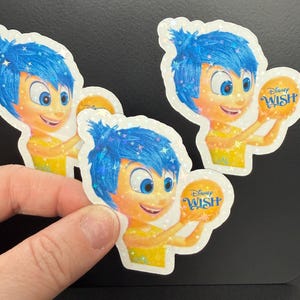 May include: Holographic stickers featuring the character Joy from the Disney movie "Inside Out." Joy has blue hair and a yellow dress, holding a golden ball with the text "Disney Wish."