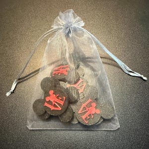 May include: A sheer, light blue drawstring bag filled with black Mickey Mouse-shaped ornaments. Each ornament has the word "LOVE" in red, with a Mickey Mouse silhouette. The bag is tied with a light blue ribbon.