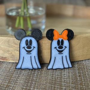 May include: Two Halloween-themed decorations. One is a blue ghost with black mouse ears and a smiling face. The other is a blue ghost with black mouse ears, an orange bow, and a smiling face. Both have a glittery finish.