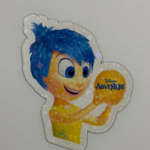 May include: A holographic sticker featuring the character Joy from the Disney movie Inside Out. Joy has blue hair and a yellow dress, holding a yellow ball with the text "Disney Adventure". The sticker has a star-shaped border.