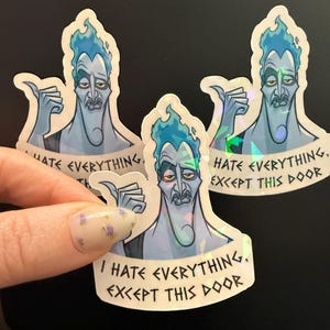 May include: Holographic stickers featuring a cartoon illustration of Hades with blue skin and flaming blue hair. The stickers have the text "I HATE EVERYTHING, EXCEPT THIS DOOR".