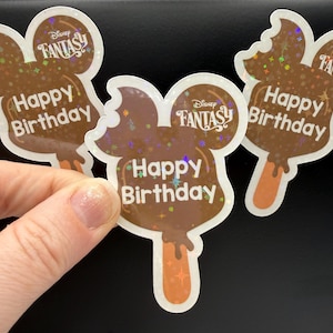 May include: Three brown Mickey Mouse-shaped stickers with the text "Happy Birthday" and "Disney Fantasy". The stickers have a popsicle stick design and a holographic glitter effect. The stickers are held by a hand.