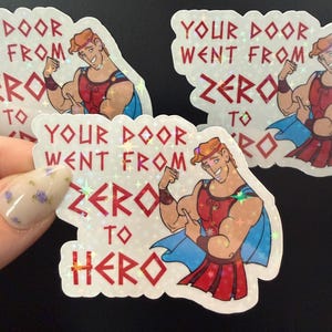 May include: A holographic sticker featuring a cartoon character with orange hair flexing a bicep, wearing a red and blue tunic. The text reads, "YOUR DOOR WENT FROM ZERO TO HERO" in red capital letters. The sticker has a shimmering, star-patterned finish.