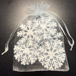 May include: A sheer, light blue organza drawstring pouch containing three white, six-pointed snowflake cutouts. The snowflakes feature intricate, symmetrical branch patterns and are visible through the semi-transparent fabric of the bag.