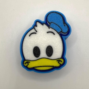 May include: A blue and white Donald Duck-shaped charm. The charm features a white textured face, black eyes, a yellow beak, and a blue hat. The charm is outlined in blue.