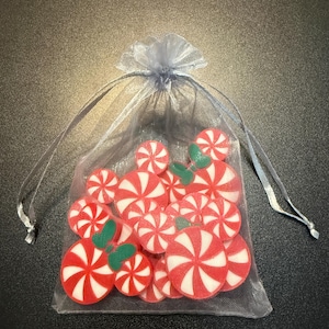 May include: A collection of red and white peppermint candy-shaped embellishments with green leaf accents, contained within a sheer, light blue drawstring bag. The candies have a spiral design. The bag is tied with a light blue ribbon.