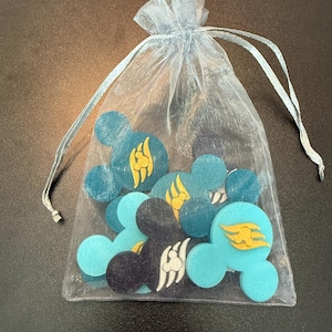 May include: A collection of teal, light blue, and black Mickey Mouse-shaped embellishments with gold and white accents, contained within a sheer, light blue drawstring bag. The embellishments feature a stylized flame design.