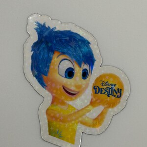 May include: A holographic sticker featuring the character Joy from the Disney movie Inside Out. Joy has blue hair and a yellow dress, holding a yellow orb with the text "Disney Destiny". The sticker has a star-shaped border.