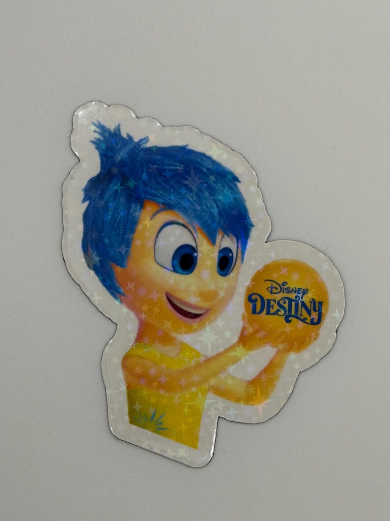 May include: A holographic sticker featuring the character Joy from the Disney movie Inside Out. Joy has blue hair and a yellow dress, holding a yellow orb with the text "Disney Destiny". The sticker has a star-shaped border.