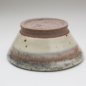 Peut inclure: Un petit bol en céramique blanche avec un glaçage moucheté brun et gris. Le bol a une base large et peu profonde et un bord légèrement évasé.