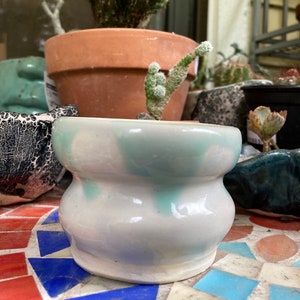 Può includere: Un piccolo vaso in ceramica bianca con una glassa blu chiaro. Il vaso ha una forma arrotondata e bulbosa con un bordo leggermente svasato. È appoggiato su una superficie di mosaico.