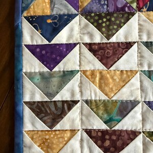 May include: Close-up of a handmade quilt with a repeating pattern of colorful triangles. The triangles are in shades of purple, gold, blue, and brown, set against a white background. The quilt is bordered with a blue fabric.