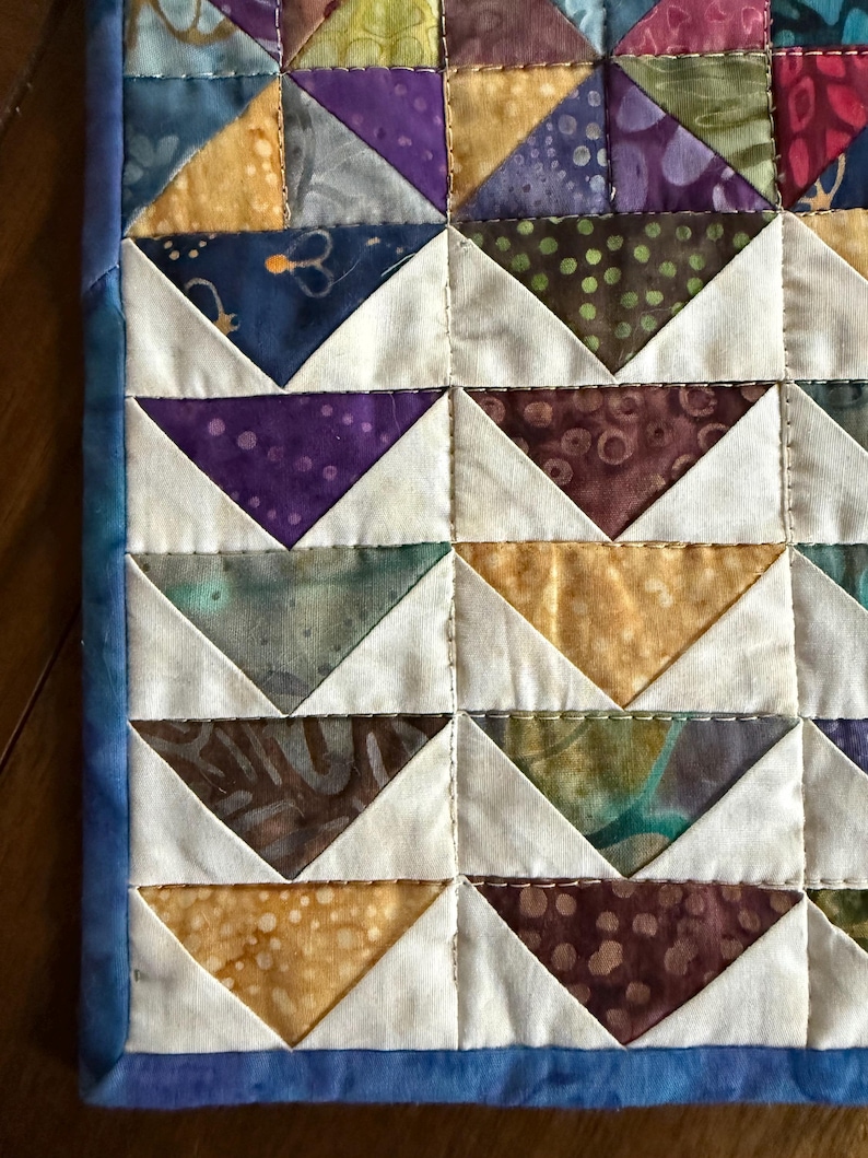 May include: Close-up of a handmade quilt with a repeating pattern of colorful triangles. The triangles are in shades of purple, gold, blue, and brown, set against a white background. The quilt is bordered with a blue fabric.