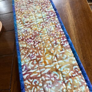 May include: A rectangular quilted table runner with a batik-style pattern in shades of yellow, orange, and purple. The runner is bordered with a blue fabric and has a quilted grid design. The runner is approximately 36 inches long.