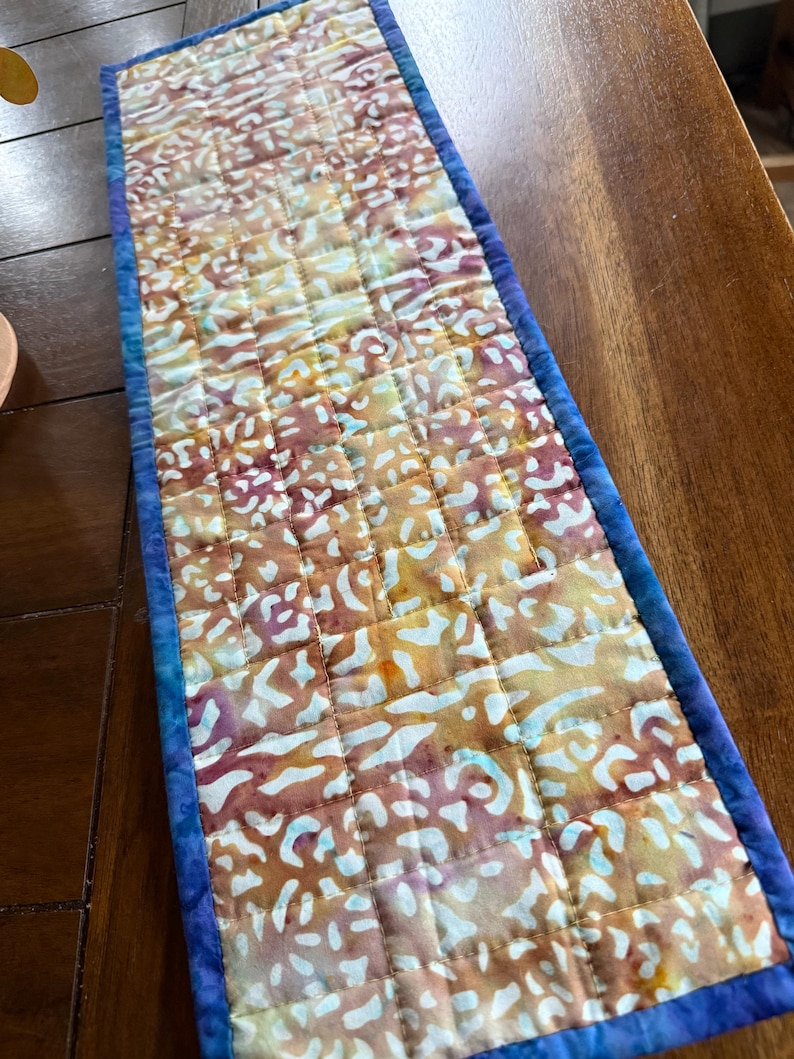 May include: A rectangular quilted table runner with a batik-style pattern in shades of yellow, orange, and purple. The runner is bordered with a blue fabric and has a quilted grid design. The runner is approximately 36 inches long.