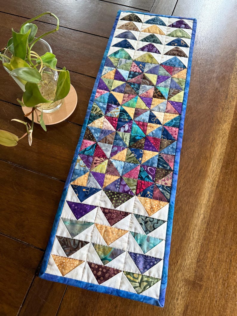 May include: A handmade, rectangular table runner with a patchwork design. The runner features a repeating pattern of colorful triangles in shades of blue, purple, green, and gold, bordered by a blue edge. The runner is approximately 12 inches wide and 36 inches long.