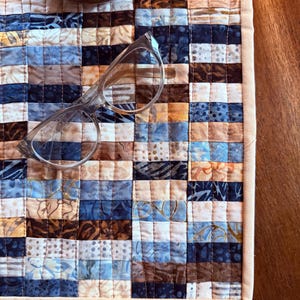 May include: A handmade, quilted table runner with a patchwork design in shades of blue, brown, and cream. Clear eyeglasses rest on the runner. A wooden coaster and a white ceramic object are in the background. The runner is bordered with a cream-colored fabric.