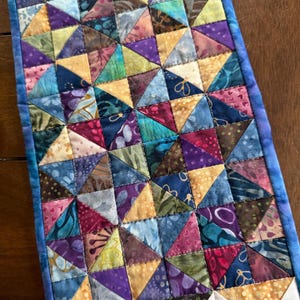 May include: A handmade quilt featuring a patchwork design of colorful triangles in shades of blue, purple, yellow, and green. The quilt has a blue border and is likely used as a table runner or decorative piece.
