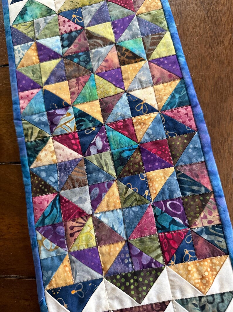 May include: A handmade quilt featuring a patchwork design of colorful triangles in shades of blue, purple, yellow, and green. The quilt has a blue border and is likely used as a table runner or decorative piece.