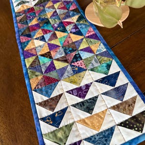 May include: A handmade, rectangular table runner with a patchwork design. The runner features a vibrant array of colorful triangles in various fabrics, including blues, purples, greens, and yellows. The edges are finished with a blue border.