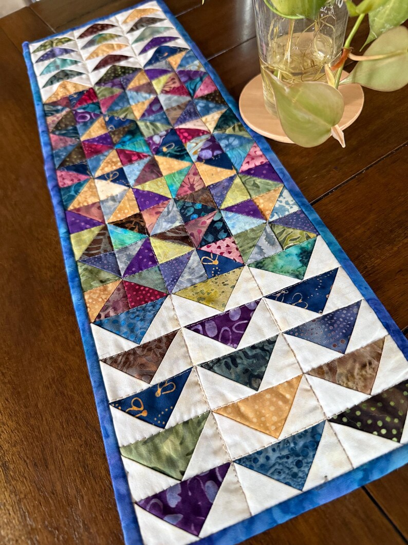 May include: A handmade, rectangular table runner with a patchwork design. The runner features a vibrant array of colorful triangles in various fabrics, including blues, purples, greens, and yellows. The edges are finished with a blue border.