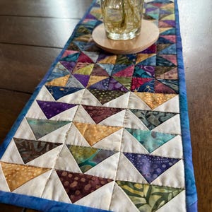 May include: A colorful, quilted table runner with a patchwork design. The runner features a repeating pattern of triangles in various colors, including blue, purple, yellow, and green. It has a blue border and is placed on a wooden surface.