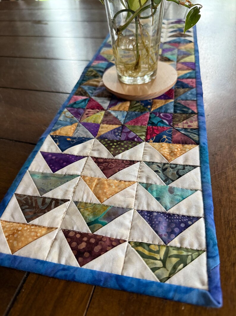 May include: A colorful, quilted table runner with a patchwork design. The runner features a repeating pattern of triangles in various colors, including blue, purple, yellow, and green. It has a blue border and is placed on a wooden surface.