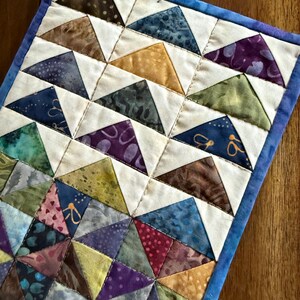 May include: A handmade quilted placemat featuring a geometric pattern of colorful triangles. The triangles are in shades of blue, green, purple, brown, and yellow, set against a white background. The placemat is bordered with a blue fabric.
