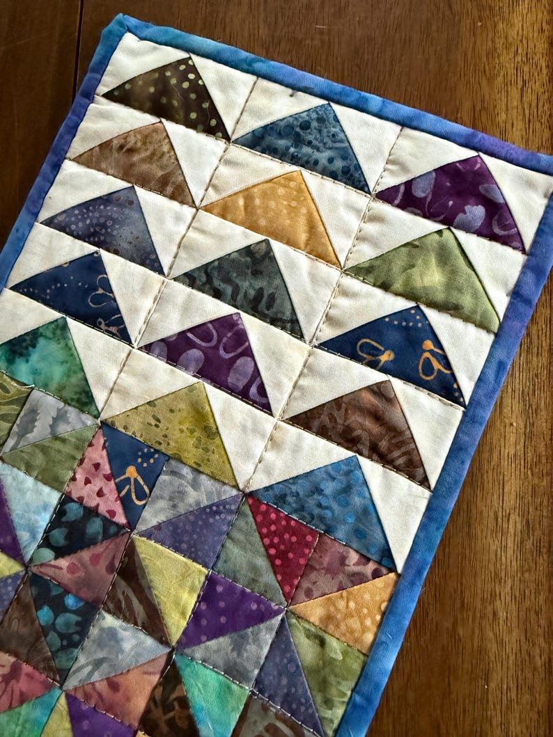 May include: A handmade quilted placemat featuring a geometric pattern of colorful triangles. The triangles are in shades of blue, green, purple, brown, and yellow, set against a white background. The placemat is bordered with a blue fabric.