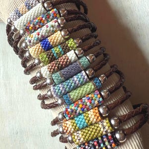 May include: A collection of beaded bracelets with brown leather straps. Each bracelet features a cylindrical beaded section in various colors, including white, blue, green, and orange. The bracelets are displayed on a neutral-colored surface.