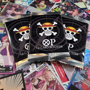 May include: Three black One Piece trading card game booster packs featuring a white skull and crossbones wearing a straw hat logo, with the text OP 12-CARD BOOSTER printed in white, resting on a scattered collection of various colorful character cards.