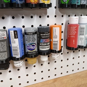 May include: A pegboard with various bottles of acrylic paint in different colors, including black, gray, blue, orange, and red. The bottles are labeled with brand names like "Rust-Oleum", "DecoArt", "REATEX", and "TRANSPARENT".