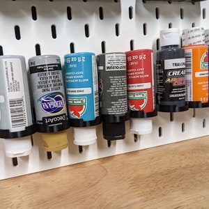 May include: A white pegboard with eight bottles of acrylic paint. The bottles are arranged in a row from left to right. The bottles are all different colors, including blue, gray, red, black, and brown. The bottles are labeled with the brand name "DecoArt" and "Apple Barrel". The bottles are all 2 fluid ounces.