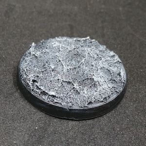 May include: A round, black plastic base with a textured, white, rocky surface. The base is designed for miniature figures and gaming.