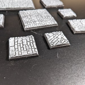May include: A set of nine grey and white textured square tiles. The tiles have a cobblestone design and are ideal for tabletop gaming.