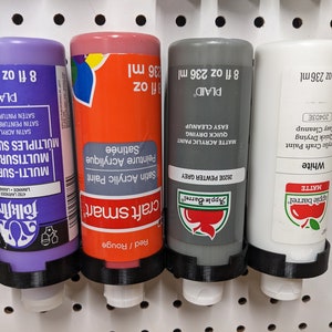 May include: Four bottles of acrylic craft paint in purple, red, gray, and white, each held in a black holder. The bottles are labeled with the brand names, paint type, and color. Each bottle contains 8 fl oz.