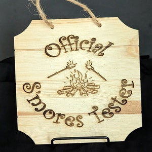 May include: A wooden sign with the text "Official Smores Tester" and a drawing of a campfire with marshmallows on sticks.