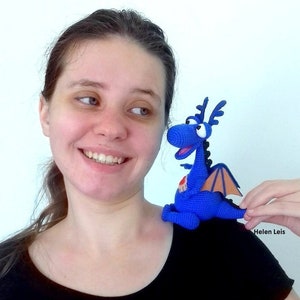 May include: A woman smiles while holding a blue crocheted dragon toy with orange wings. The dragon has a red heart on its chest and large, round eyes. The woman is wearing a black t-shirt with a silver design.